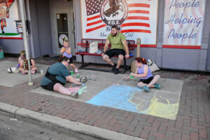 Family chalk painting the sidewalk in front of the New Lexington Eagles on Main Street during the 2026 Chalk It Up to Summer event for kids and families.