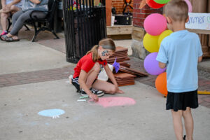 Face-painted girl chalks the sidewalk on Main Street in New Lexington during the 2026 Chalk It Up to Summer event for kids and families.