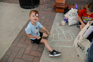 Face-painted boy points to his chalk drawing on Main Street in New Lexington during the 2026 Chalk It Up to Summer event for kids and families.
